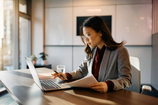 Asian female executive working on laptop while going through business reports in the office.