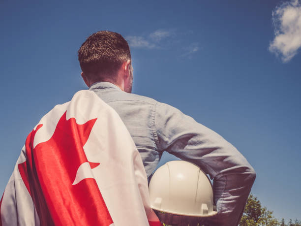 Young engineer, white hardhat and Canadian Flag in the park against the backdrop of green trees and the setting sun, looking into the distance. Close-up. Concept of labor and employment