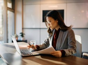 Asian female executive working on laptop while going through business reports in the office.