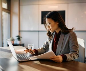 Asian female executive working on laptop while going through business reports in the office.