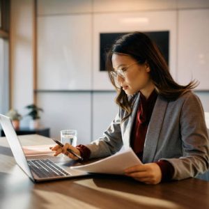 Asian female executive working on laptop while going through business reports in the office.