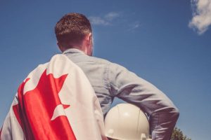 Young engineer, white hardhat and Canadian Flag in the park against the backdrop of green trees and the setting sun, looking into the distance. Close-up. Concept of labor and employment