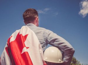 Young engineer, white hardhat and Canadian Flag in the park against the backdrop of green trees and the setting sun, looking into the distance. Close-up. Concept of labor and employment