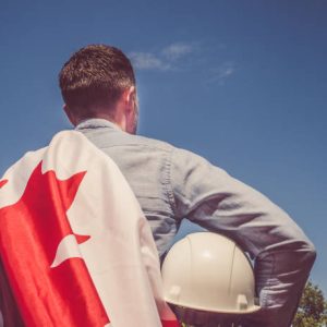 Young engineer, white hardhat and Canadian Flag in the park against the backdrop of green trees and the setting sun, looking into the distance. Close-up. Concept of labor and employment