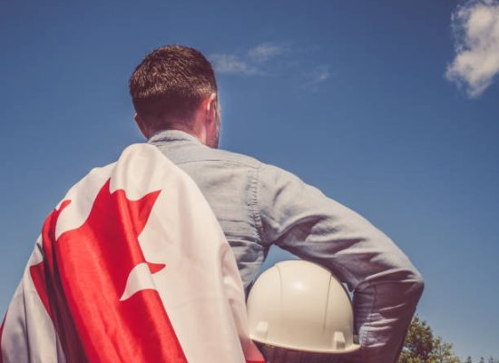 Young engineer, white hardhat and Canadian Flag in the park against the backdrop of green trees and the setting sun, looking into the distance. Close-up. Concept of labor and employment