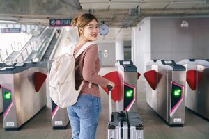 Asian woman traveler smiling while pulling suitcase and passing automatic gate at modern train station. Concept of solo travel, public transportation, smooth transit, urban mobility and confident travel lifestyle.