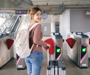 Asian woman traveler smiling while pulling suitcase and passing automatic gate at modern train station. Concept of solo travel, public transportation, smooth transit, urban mobility and confident travel lifestyle.