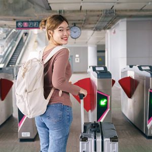 Asian woman traveler smiling while pulling suitcase and passing automatic gate at modern train station. Concept of solo travel, public transportation, smooth transit, urban mobility and confident travel lifestyle.