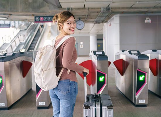 Asian woman traveler smiling while pulling suitcase and passing automatic gate at modern train station. Concept of solo travel, public transportation, smooth transit, urban mobility and confident travel lifestyle.
